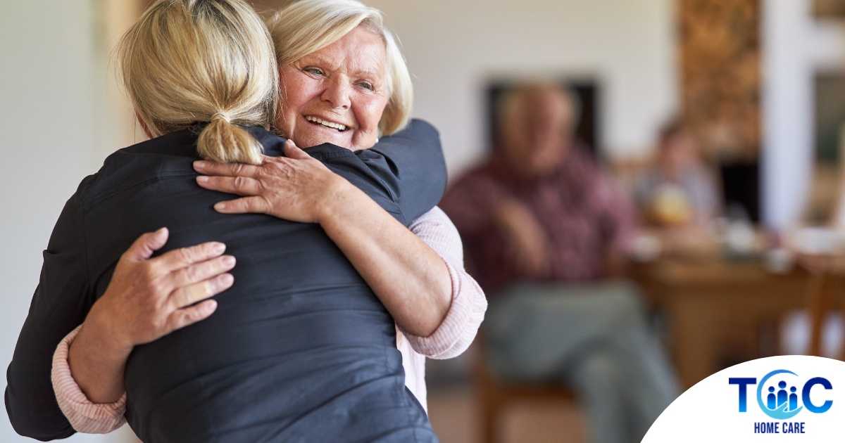 An older woman smiles as a younger woman visits her and hugs her, showing the effect that acts of kindness can have on senior loved ones.